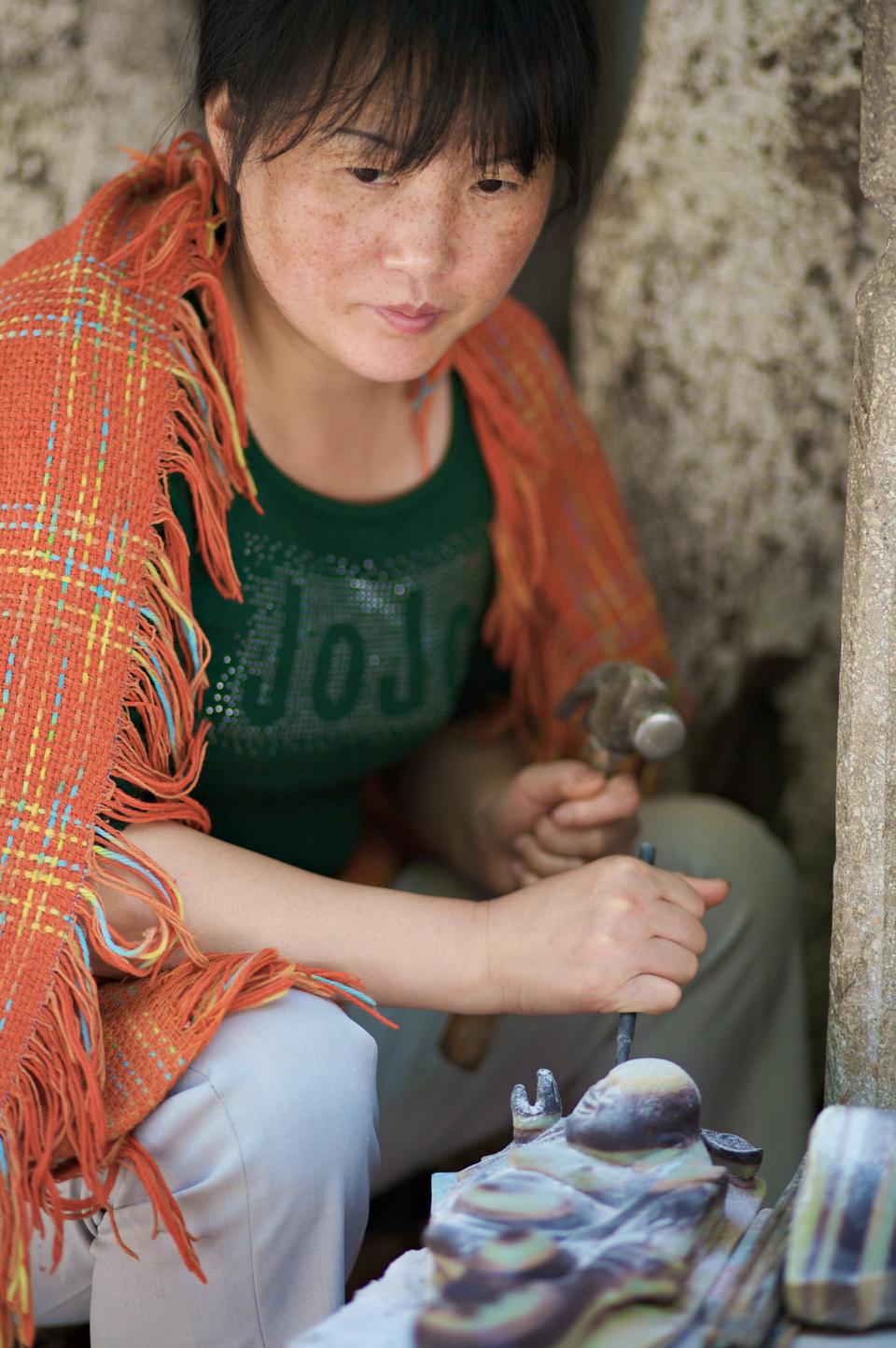 Many people in China make their living as artisans and salesmans in street stalls. This woman pauses for a moment to listen to a passerby as she is working on her Buddhas.
