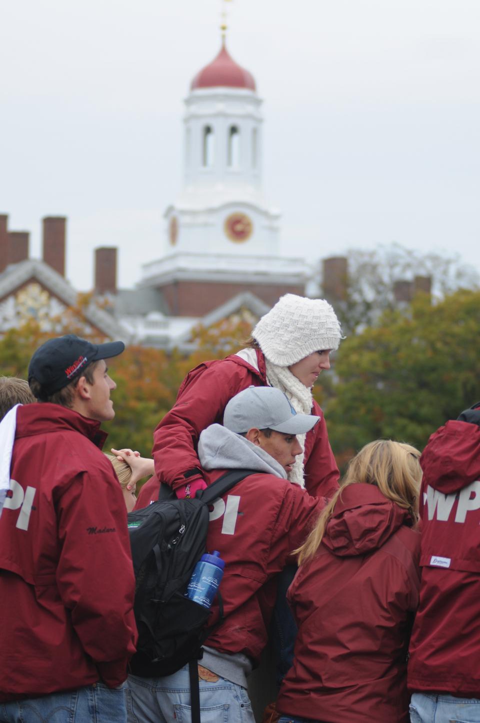 A crew team member perches on a bridge railing, supported by another crew team member. Intense onlookers crowded the various Charles River bridges this weekend.