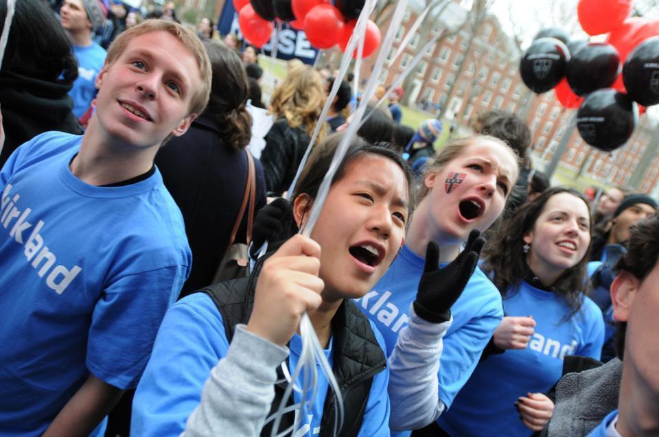 Upperclassmen run through the Yard to deliver results to eager freshmen. Near the John Harvard statue, enthusiastic Kirkland House residents show their spirit with cheers, balloons, and temporary face tattoos.