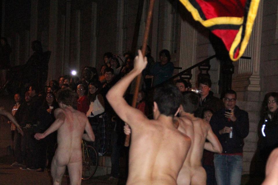 A Primal Scream participant carries the Adams House flag past a crowd in front of the John Harvard Statue during the first few minutes of Finals Period, joining, among others, skateboarders and a student in a hamburger suit.