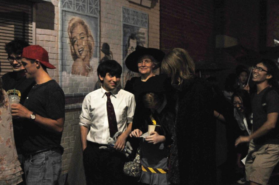 Two friends wait on line for the new Harry Potter movie while another reads. Behind them, a woman dressed as a witch waits to see the film.