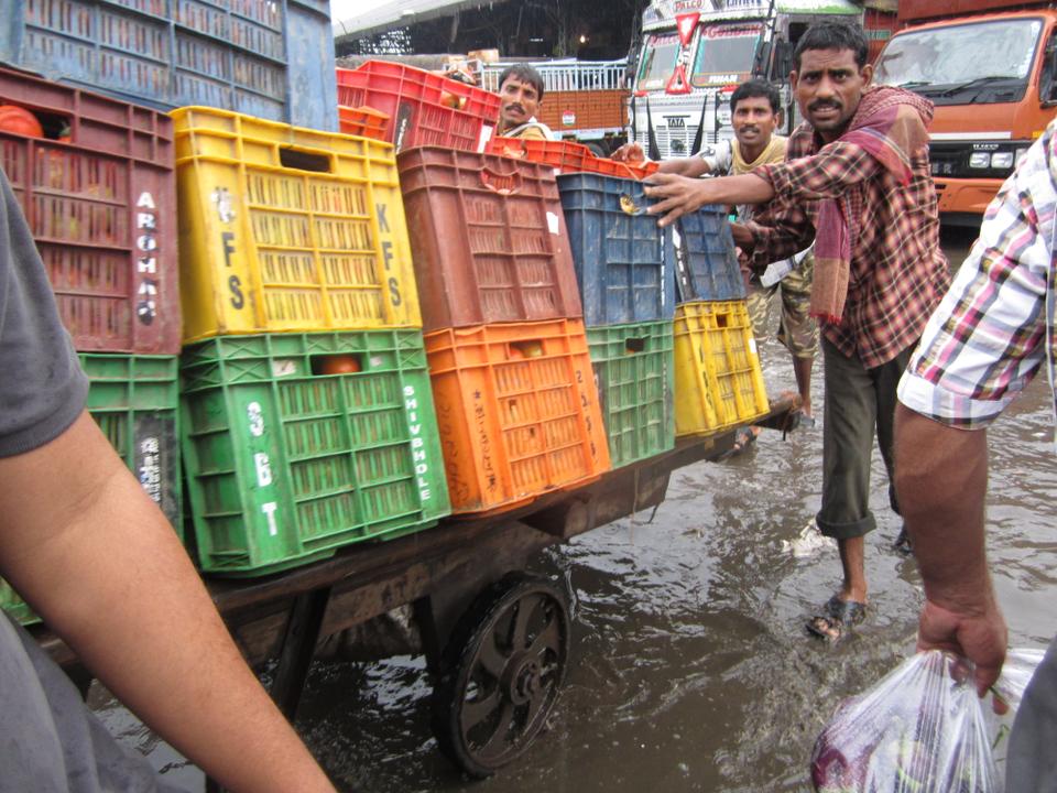 Men push a handcart stacked high with tomato crates through the monsoon-flooded parking lot at Azadpur Mandi.