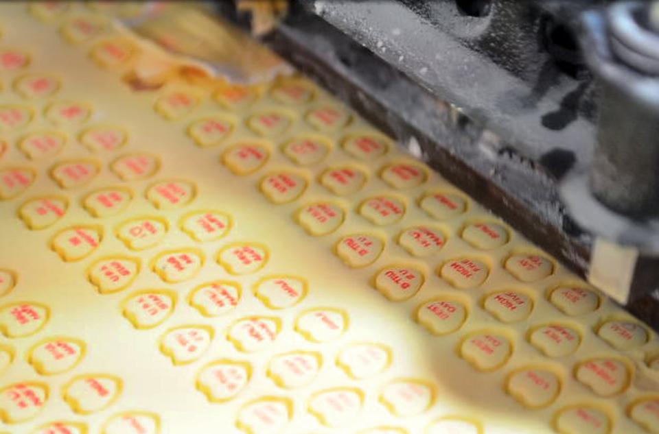 A roll of banana-flavored wafer dough is rolled through a series of rollers that prints the typical "Sweethearts" phrases on the dough and stamps the dough into heart-shaped pieces befoer they are separated and baked.