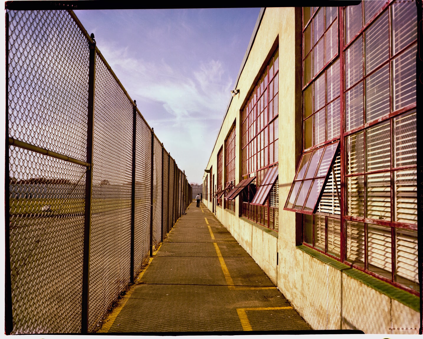 Men walk between a chain link fence and one of the buildings in Walpole Prison.